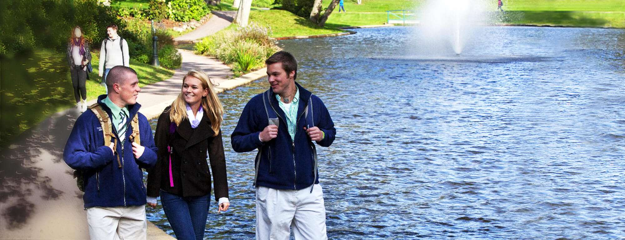 two young men and a young woman walking and talking on a path near a pond with a fountain