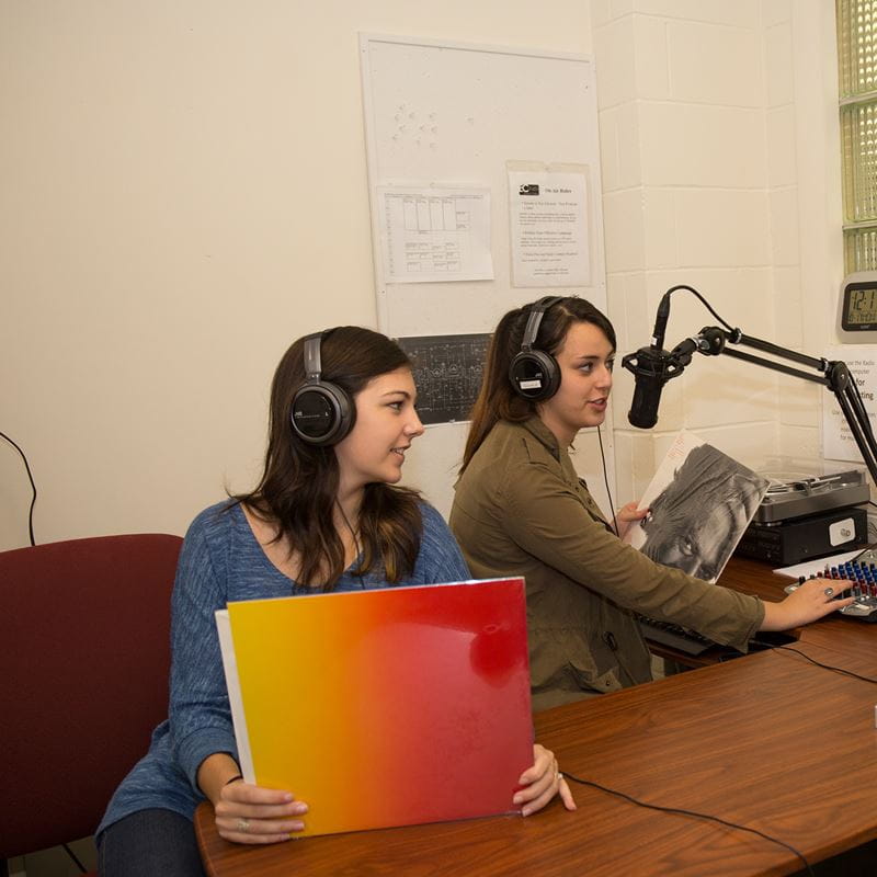 two students working in radio station with headphones on one holding a record case