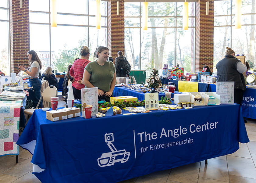 Student vendor smiling behind a blue table with baked goods and festive decorations in a sunlit hall.