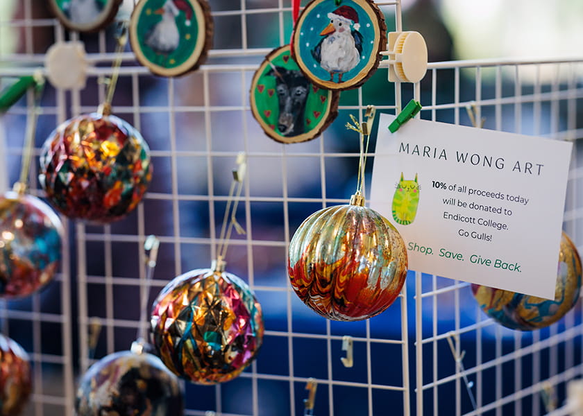 Handcrafted glass ornaments and painted wooden discs hanging on a white grid display next to a sign for "Maria Wong Art".