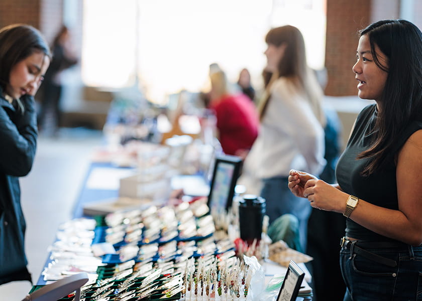 Vendor standing at her booth talking to a customer browsing trays of handmade earrings.