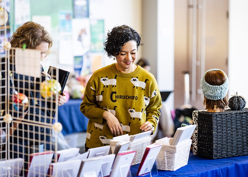 Woman in a green sweater with cat patterns looking down at greeting card displays on a table at a craft market.