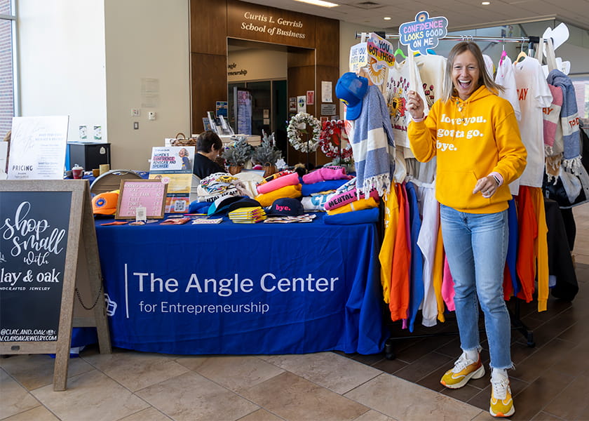 Smiling woman in a yellow hoodie holding up a sign beside apparel racks at "The Angle Center for Entrepreneurship" booth.
