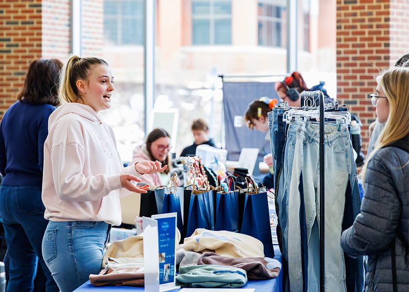 Young woman in a pink hoodie gesturing while speaking with a customer at a clothing vendor booth.