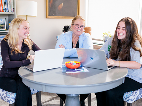 Two students and a woman with glasses sit around a round table, laughing together while using laptops in an office