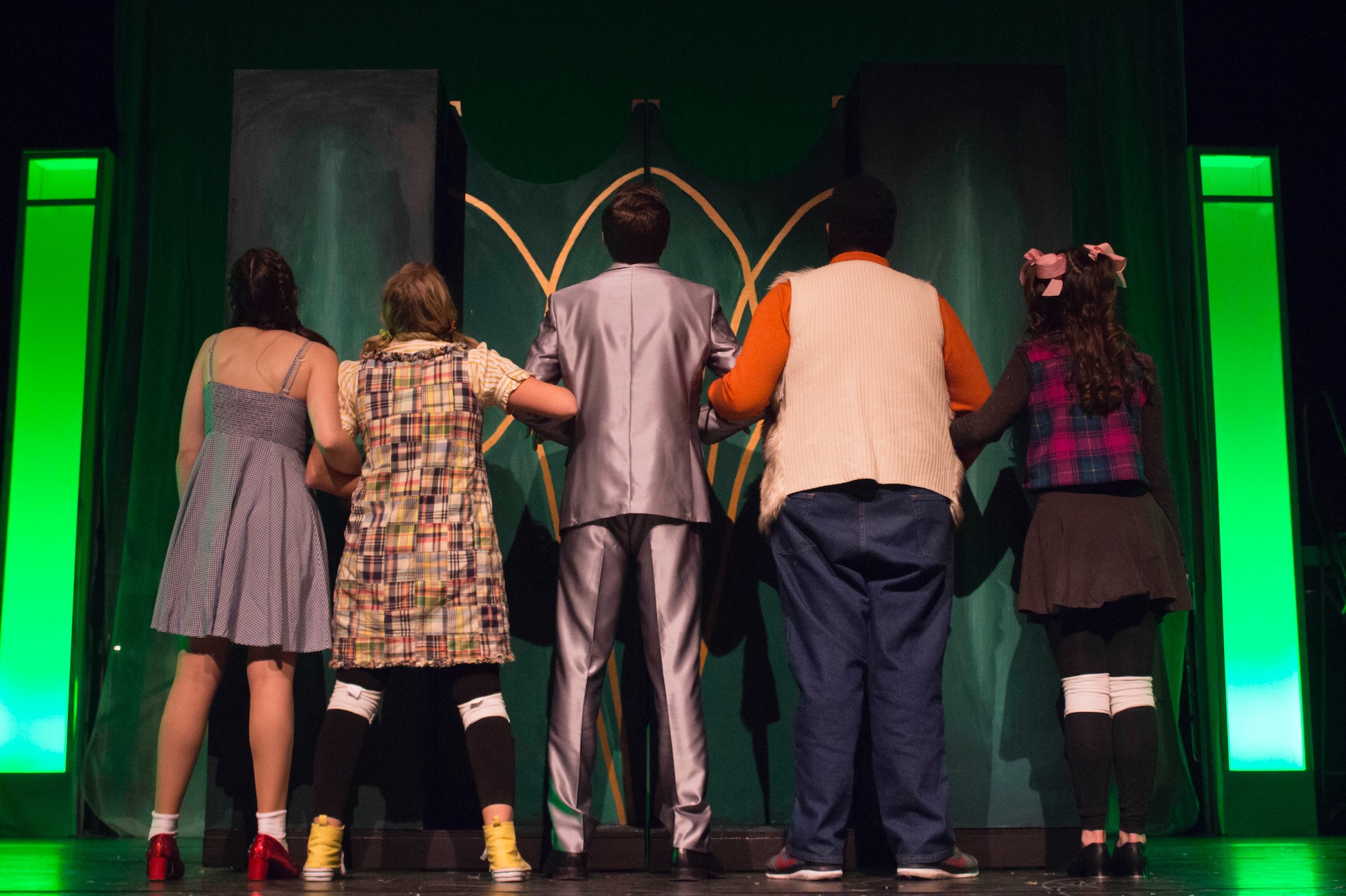 main cast of wizard of oz play characters stand facing the back of the stage with arms linked