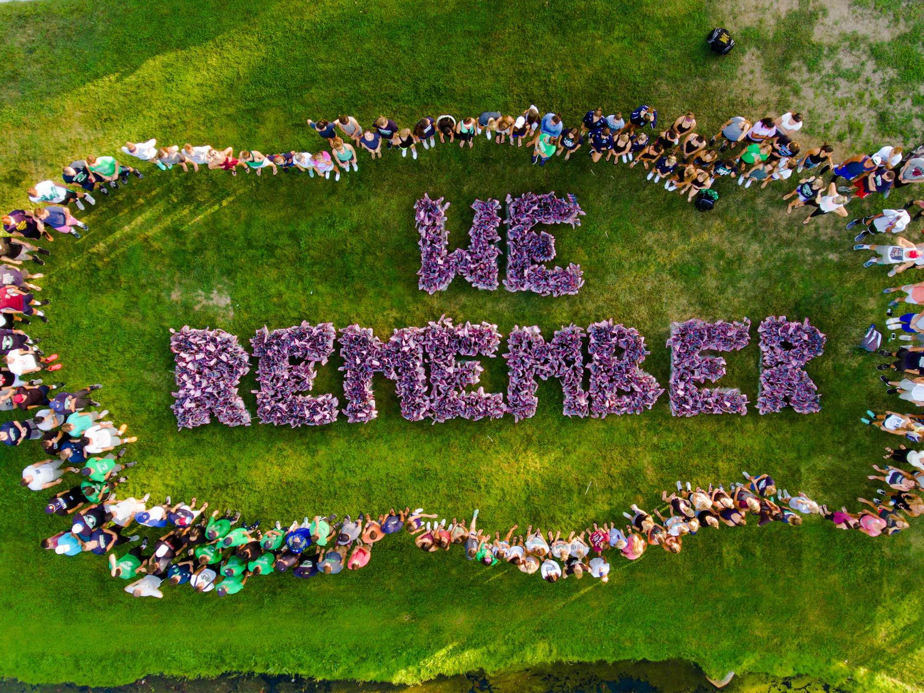 a top-down view of many people standing around the words 'we remember' spelled out with US flags