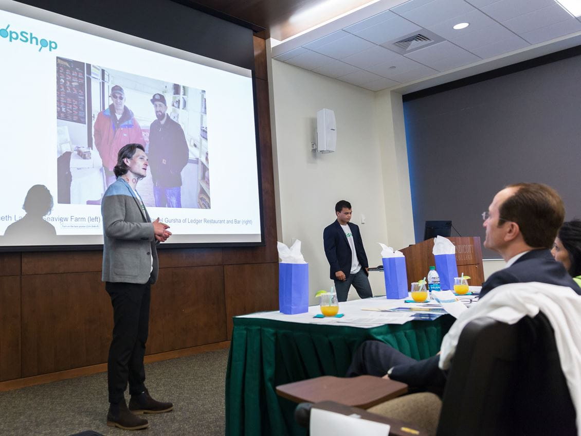 A man standing in front of a projector pitches his business idea to a panel of judges