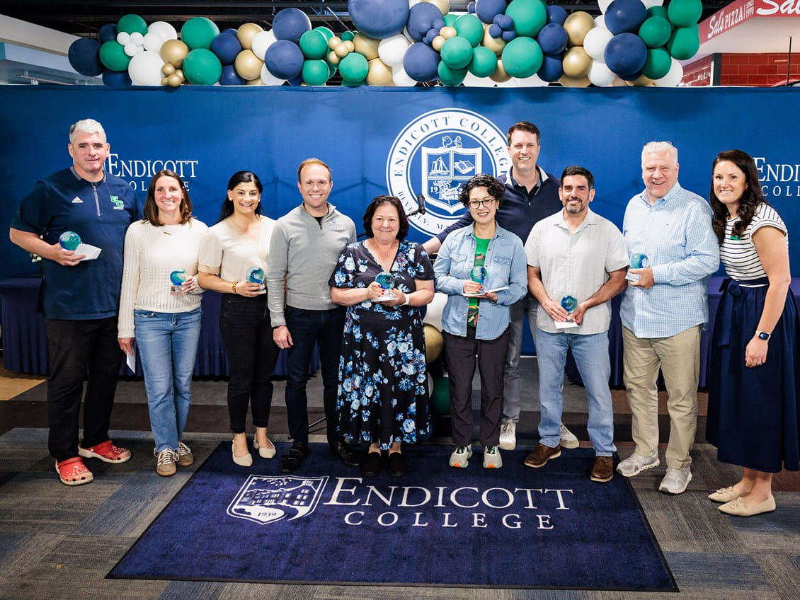 Ten smiling people pose holding awards at an Endicott College event, standing in front of a blue banner and a green balloon arch.