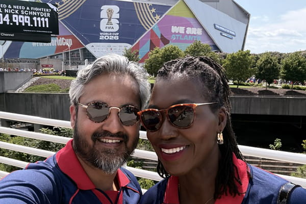 Smiling man and woman in red shirts and sunglasses pose outside a FIFA World Cup 2026 venue in Atlanta.