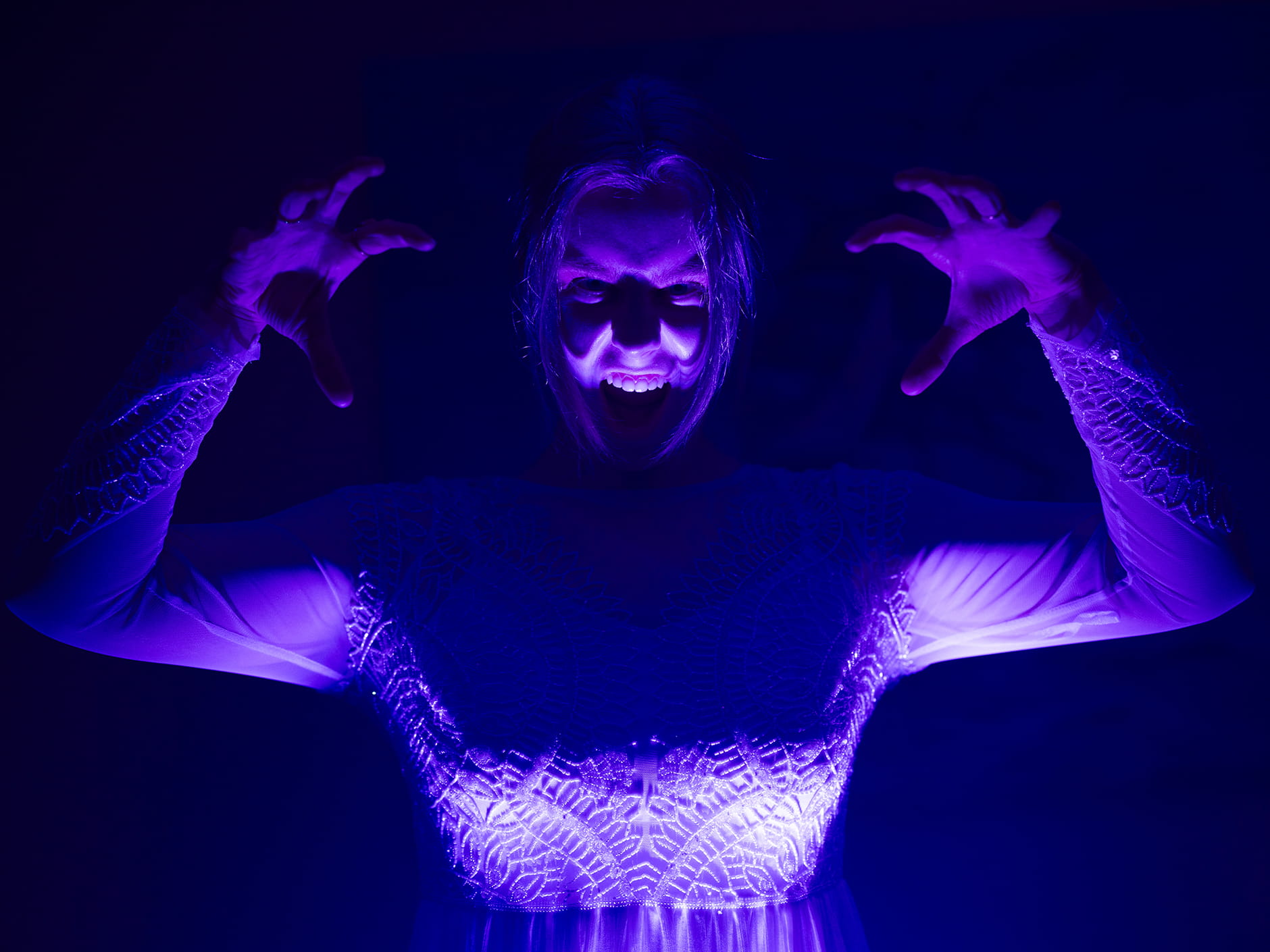 A person dressed in white raises their hands in a spooky pose, their face illuminated by eerie purple lighting against a dark background.
