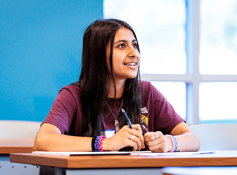 A smiling young woman with long dark hair sits at a desk, holding a pen over a notebook. She wears a maroon t-shirt and colorful bracelets while looking forward.