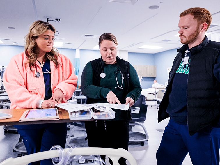 Three nursing students in a simulation lab review medical charts and a tablet at a workstation. One points to a document while others look on intently.