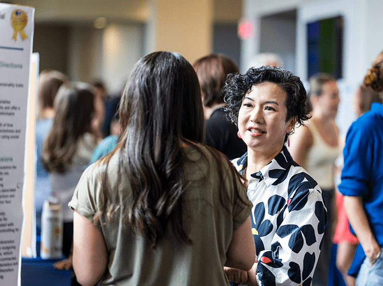 An academic professional in a floral shirt speaks with a student during a poster presentation session in a crowded, brightly lit campus hallway.