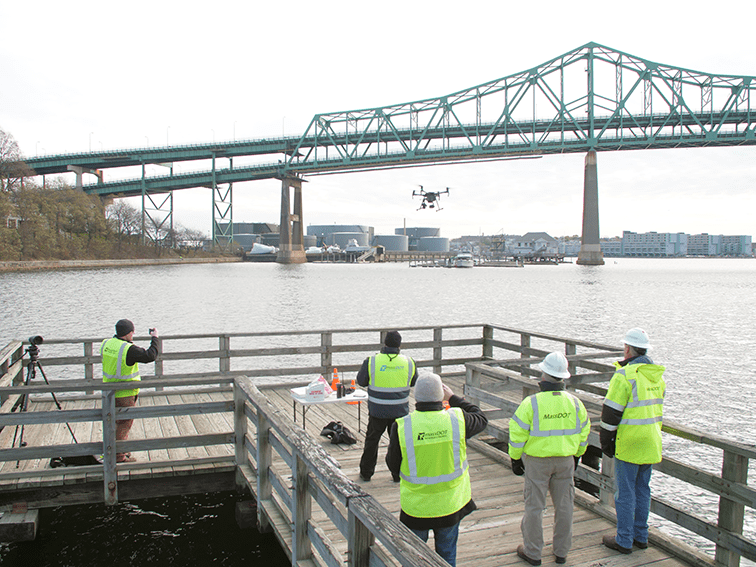 Five MassDOT workers in high-visibility vests stand on a wooden pier, monitoring a drone flying over a river toward a large green steel truss bridge in the background.