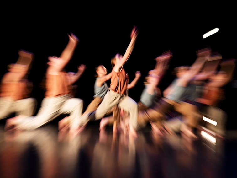 A group of dancers performing an expressive, energetic routine on a dark stage with blurred motion highlights as they reach upwards.