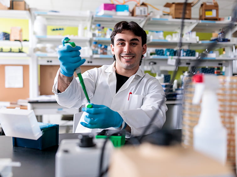 A smiling male student in a lab coat and blue gloves using a micropipette at a lab bench.