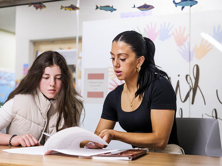 A counselor or tutor sitting at a desk with a student, pointing at open documents in a notebook or folder.