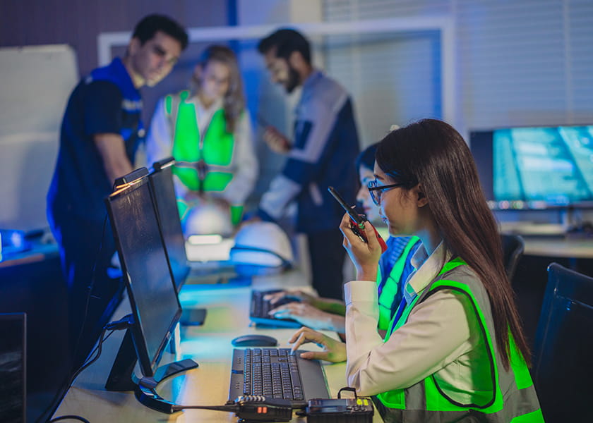 A woman in a high-visibility safety vest speaks into a walkie-talkie while working at a computer in a dim command center with colleagues collaborating in the background.