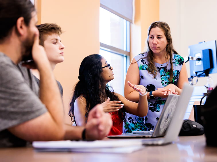 A student gesturing with her hands while speaking to a standing instructor during a group discussion in a classroom.