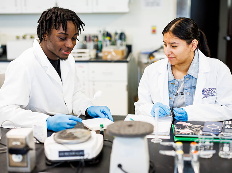 Two science students in lab coats and gloves conducting an experiment at a lab workstation.