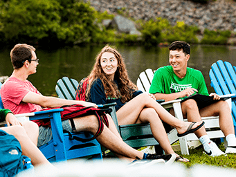 Three college students sit in Adirondack chairs by a pond, smiling and talking on a sunny day.