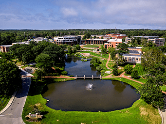 Aerial view of a scenic college campus with a bridge spanning a two-part pond with a fountain, surrounded by buildings and a large forest.