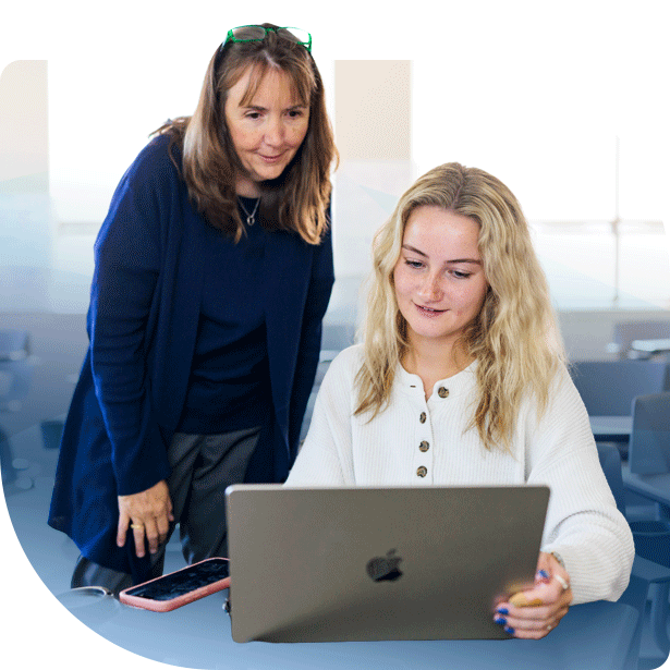 A woman with green glasses and a younger woman with blonde hair laugh together while looking at a laptop screen in a bright, indoor classroom or office setting.