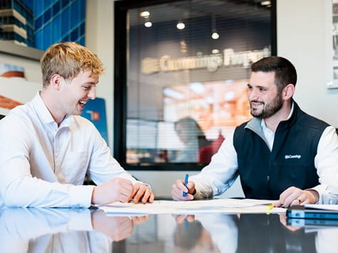Two professional men sitting at a glass table reviewing plans and chatting in a bright office space.