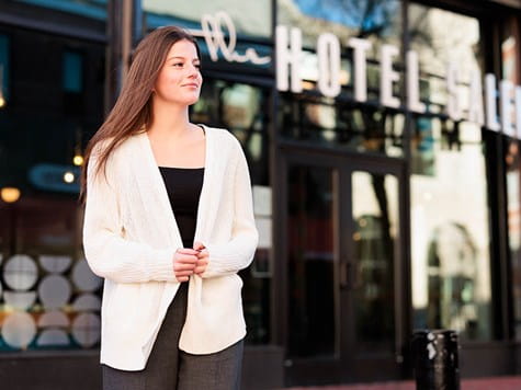 A young woman wearing a cream cardigan stands outside, looking off-camera in front of a hotel entrance.