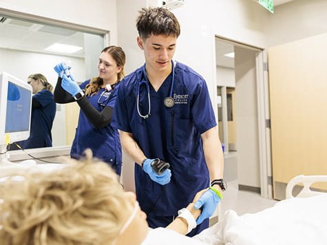 A male nursing student wearing blue scrubs scans a patient's wristband while a classmate works in the background.