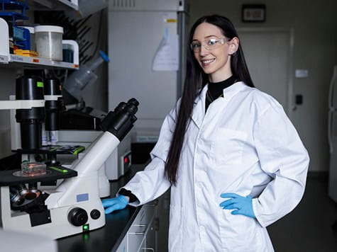A female student in a lab coat and safety glasses standing proudly next to a lab microscope.