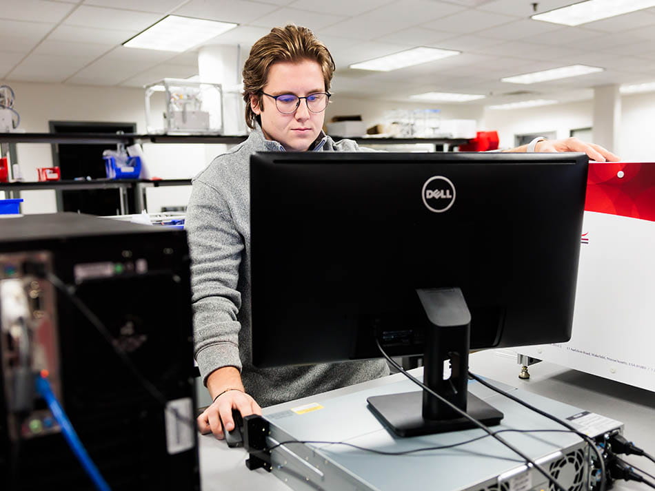 A male student wearing glasses standing in a tech lab and focusing on a large Dell monitor.