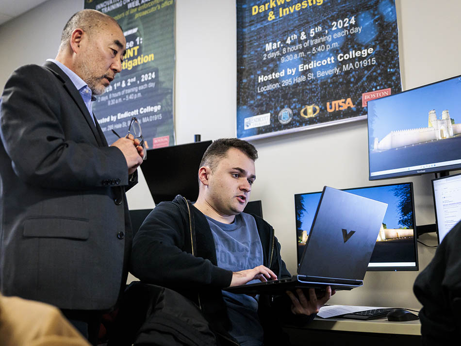 A male student with a laptop discussing work with an older male instructor standing next to him.