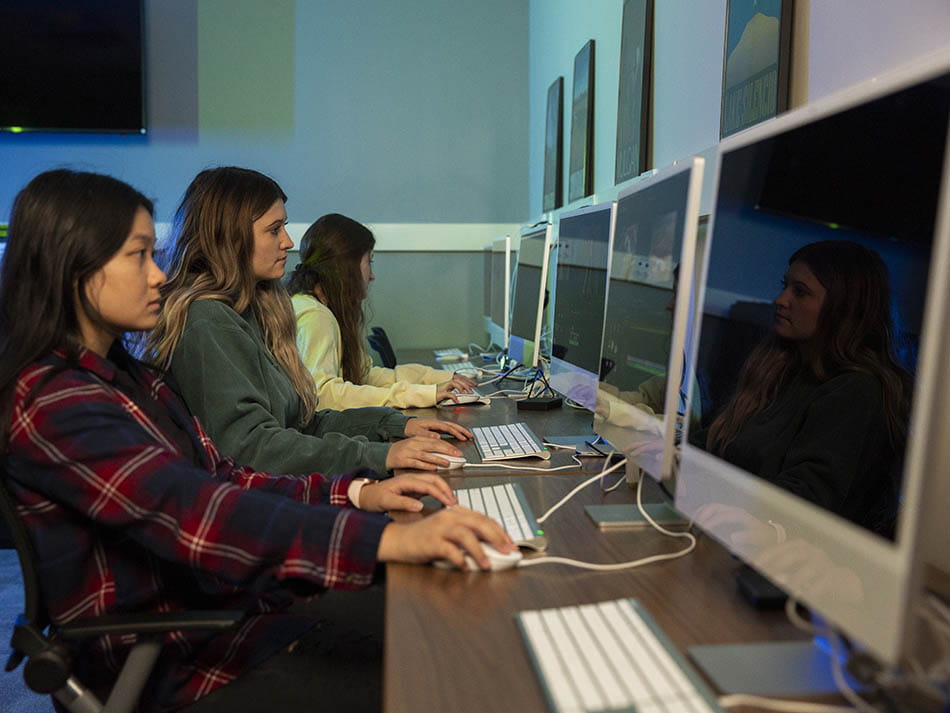 A line of students sitting side-by-side at a long desk, each working on an iMac computer.