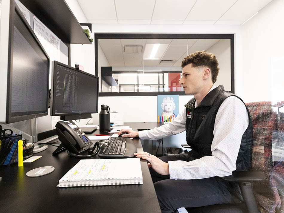 A young man in a vest sitting at an office desk working across dual computer monitors.