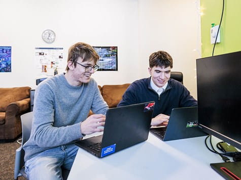 Two male students sitting at a table together, smiling and collaborating on their laptops.
