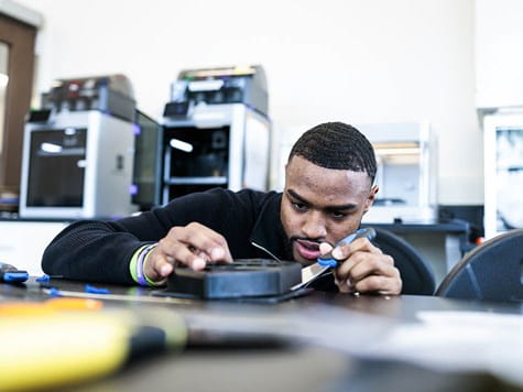 A male student carefully using a scraper tool on a 3D-printed part in an engineering lab.