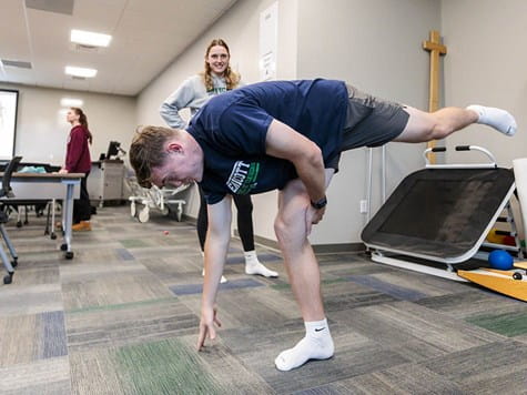 A young man balances on one leg reaching for the floor while a female instructor observes in a clinic setup.