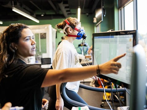 A woman wearing an oxygen mask on a treadmill while an instructor points at a screen tracking metrics.