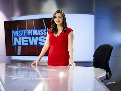 A female news anchor in a red dress standing behind a newsroom desk with "Western Mass News" displayed behind her.