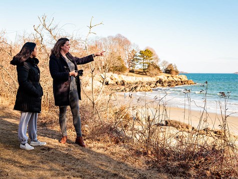 A woman pointing out toward a coastal shoreline while standing outdoors with a female student.