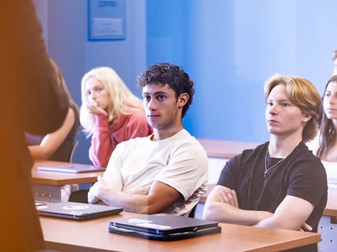 Students sitting at wooden desks in a classroom, attentively watching a lecture.