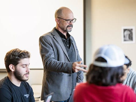 A professor wearing glasses and a blazer gesturing while leading a classroom discussion with students.