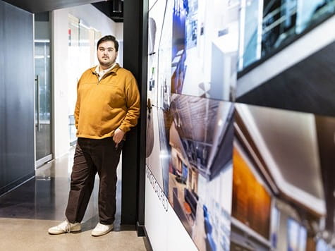 A young man leaning against a wall displaying photos of interior design projects.