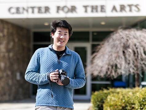 A smiling male student holding a DSLR camera outside a building labeled Center for the Arts.