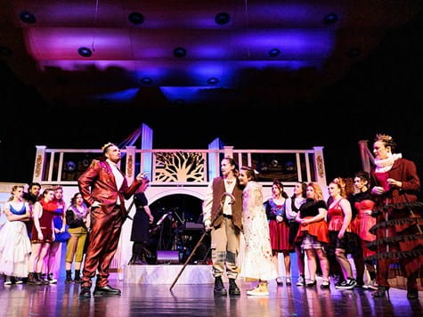Actors in elaborate costumes performing a theatrical play on a brightly lit stage set.